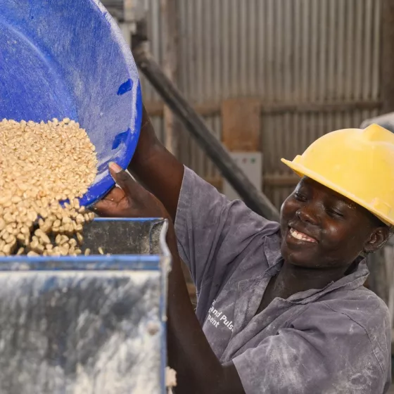 A woman worker at Awelo Millers and Packers Investment Ltd in the Oyam District, Uganda, smiles while pouring grain into a milling machine.